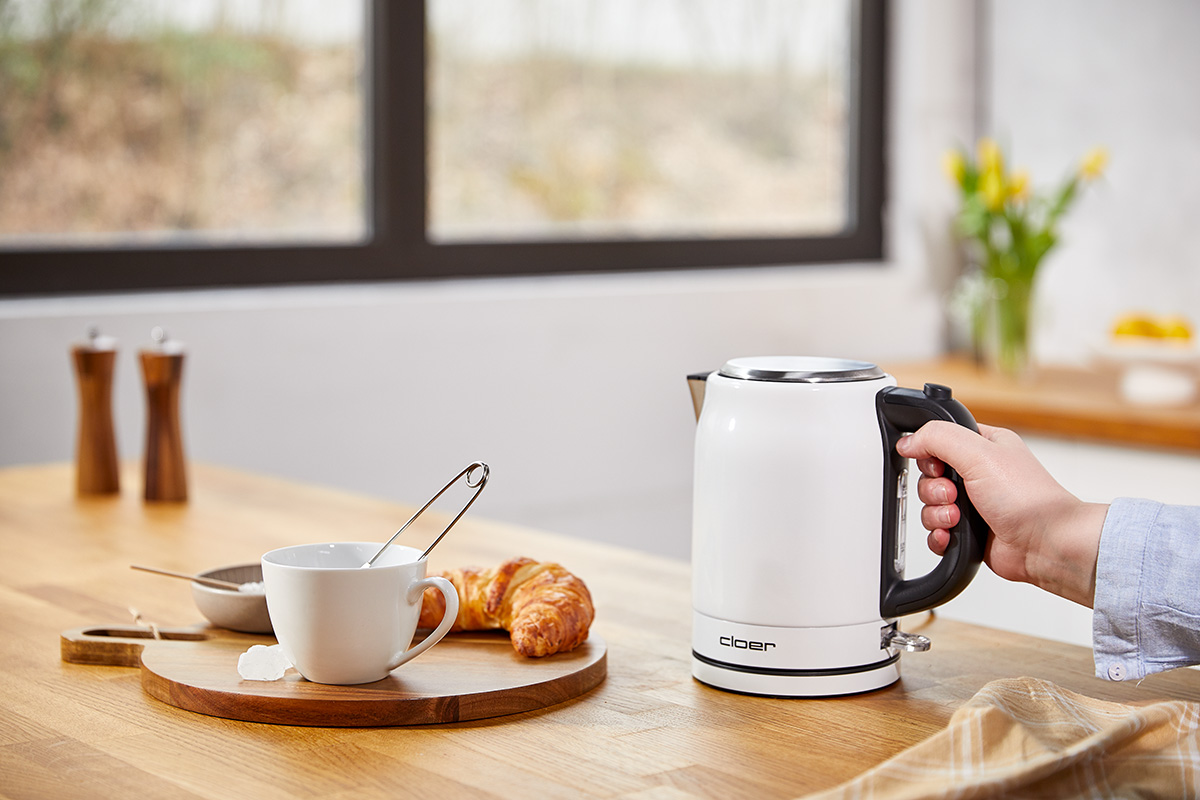 Das Bild zeigt wie jemand den Wasserkocher am Griff hält mit einer Teetasse und einem Crossaint auf einem Brettchen.