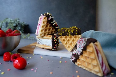 Das Bild zeigt vier Eiswaffel-Sandwiches dekoriert mit Schokolade und Zuckerperlen auf einem Holzbrett, daneben liegen Erdbeeren.