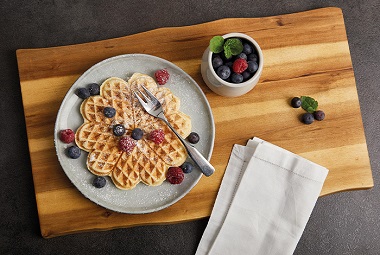 Das Bild zeigt eine Herzwaffel auf einem grauen Teller dekoriert mit Beeren, daneben ein Töpfchen mit Heidelbeeren, beides auf einem Holzbrett. 