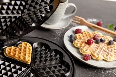 Das Bild zeigt ein halb geöffnetes Cloer Waffeleisen mit einem fertigen Waffelherz, daneben steht ein Teller mit einer Herzwaffel und Holzgabel, daneben eine Tasse mit Kaffee.