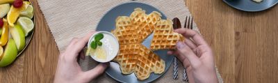 Das Bild zeigt eine Buttermilchwaffel auf einem grauen Teller, darüber eine linke Hand hält ein Töpfchen mit Zitronencreme dekoriert mit Zitrone und Minze, die rechte Hand hält ein einzelnes Waffelherz. Daneben steht ein Obstkorb mit Zitrusfrüchten.