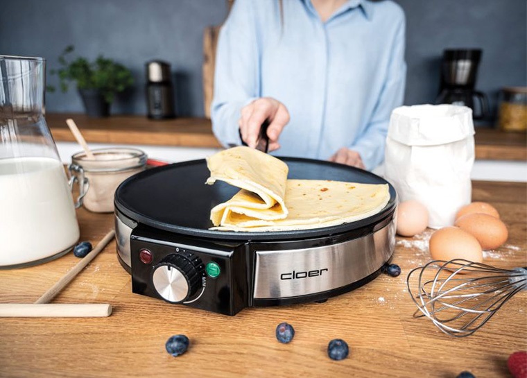 Das Bild zeigt den Crêpes Maker Cloer-6630 und eine Hand, die mit einem Wender, einen Crêpes auf der Backfläche faltet.