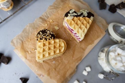 Das Bild zeigt zwei Eiswaffelsandwiches auf Backpapier mit Schokolade, Mandelblättchen und gehackten Nüssen verziert, daneben steht ein Glas mit Mini-Marshmallows.