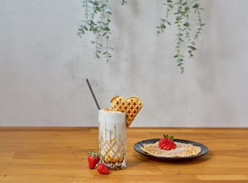 Das Bild zeigt ein Glas mit Eiskaffee, daneben steht ein Teller mit einer Waffel und Erdbeeren. 